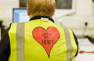Worker sitting facing computer wearing yellow hi vis jacket with 'safety at heart' written on back
