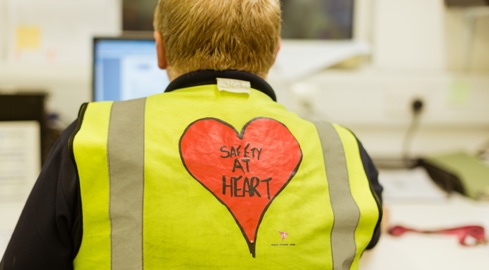 Worker sitting facing computer wearing yellow hi vis jacket with 'safety at heart' written on back