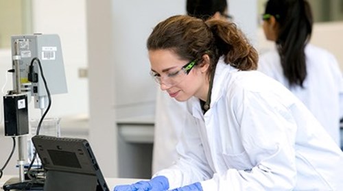 Scientist wearing white lab coat protective goggles and blue plastic gloves looks at testing machine
