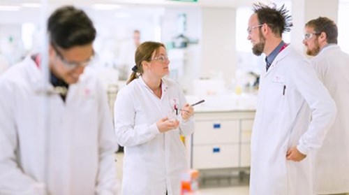 Four scientists wearing protective goggles and white lab-coats working in a Scholl lab