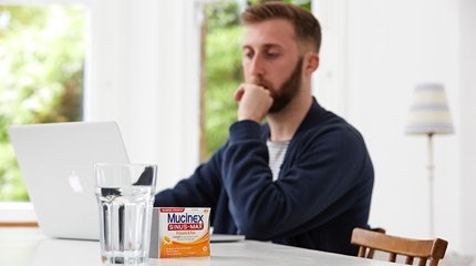 Man with pensive expression sitting at table looking at MacBook with glass of water and box of Mucinex tablets