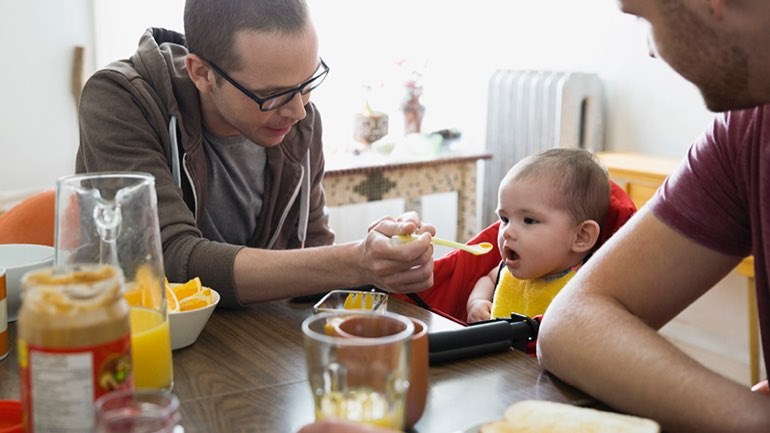 Parent wearing glasses feeds baby in high chair with yellow spoon while other adult sits next to baby