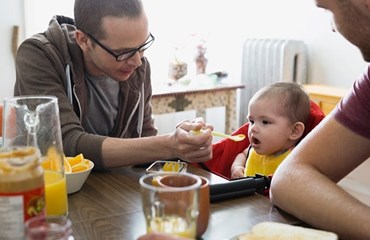 Parent wearing glasses feeds baby in high chair with yellow spoon while other adult sits next to baby