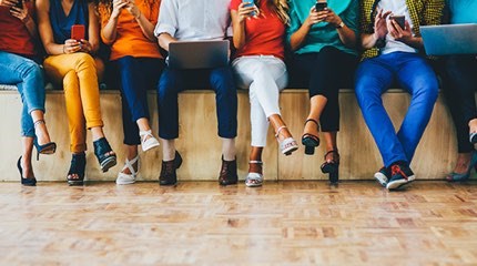 Eight people sitting on the edge of a stage looking at their phones and one in the middle looking at laptop