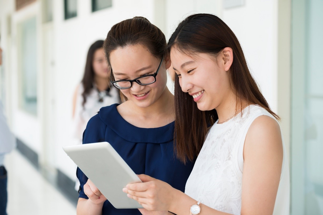 Two women, standing in a corridor, smiling and looking at an iPad 