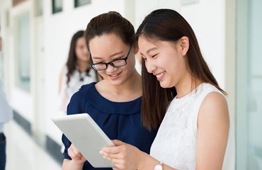 Two women, standing in a corridor, smiling and looking at an iPad 
