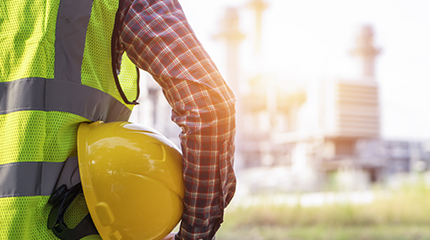 Worker looking at factory wearing hi vis jacket and holding yellow protective helmet