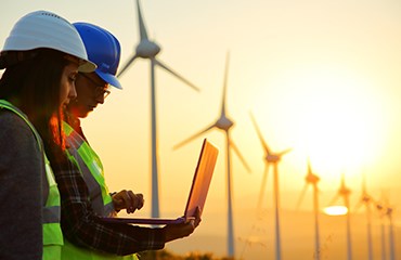 Two wind farm workers stand looking at laptop in front of wind turbines