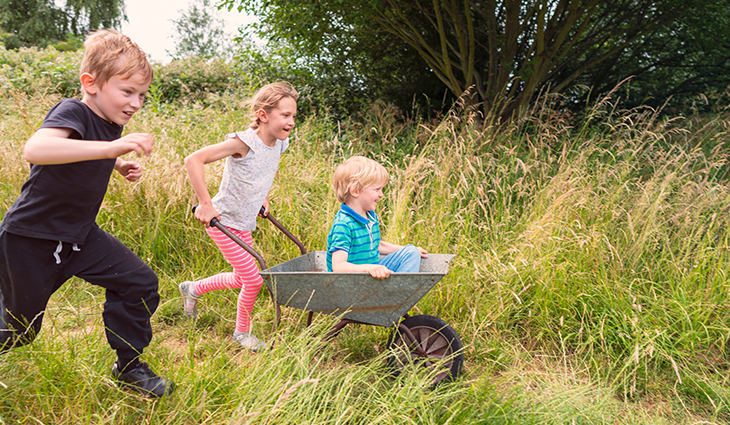 Two children running through a grassy field pushing younger child in wheelbarrow