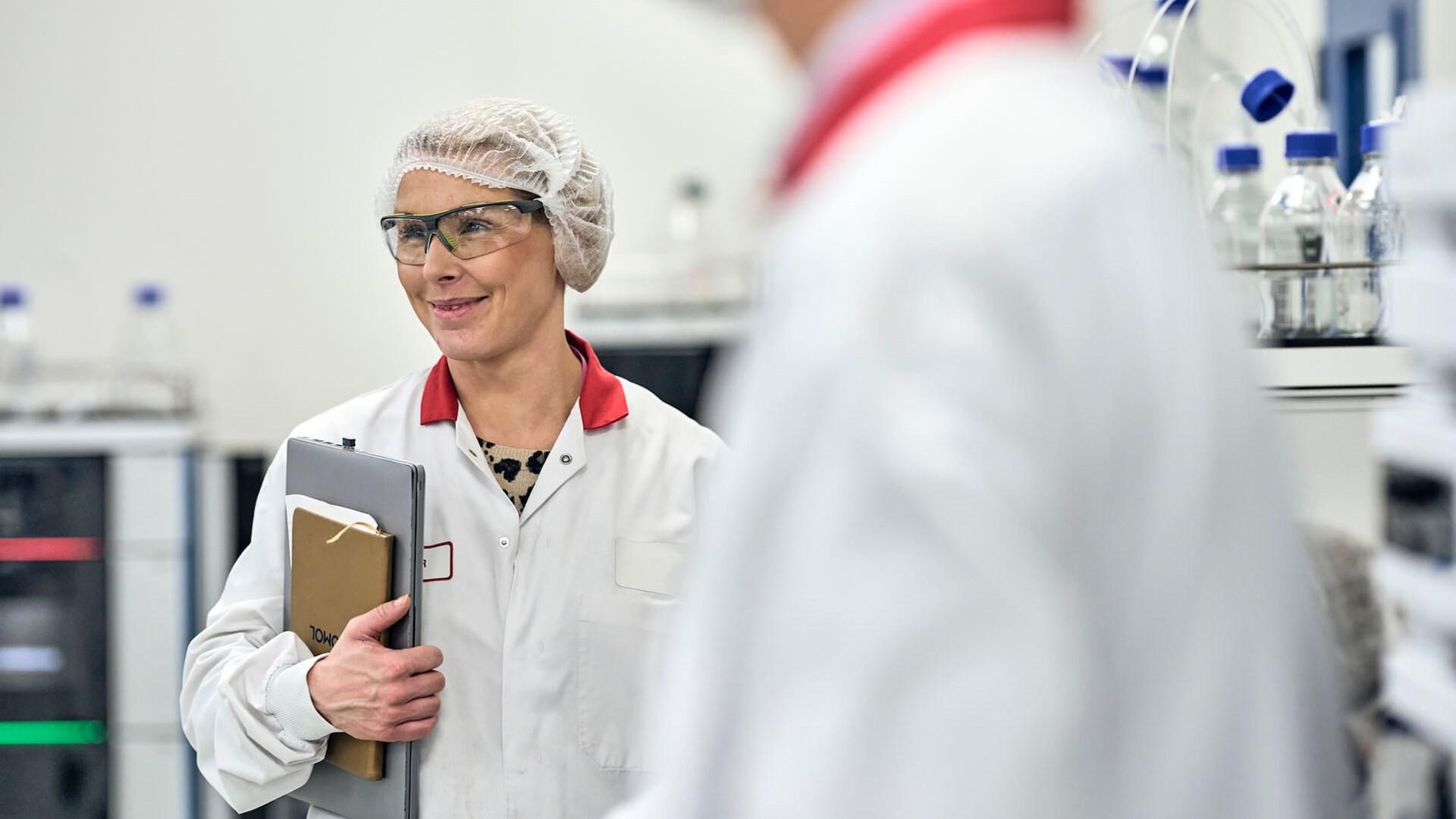 Two Reckitt employees working in a lab, wearing hairnets, safety glasses and a lab coat. One carries a clipboard and notebook.