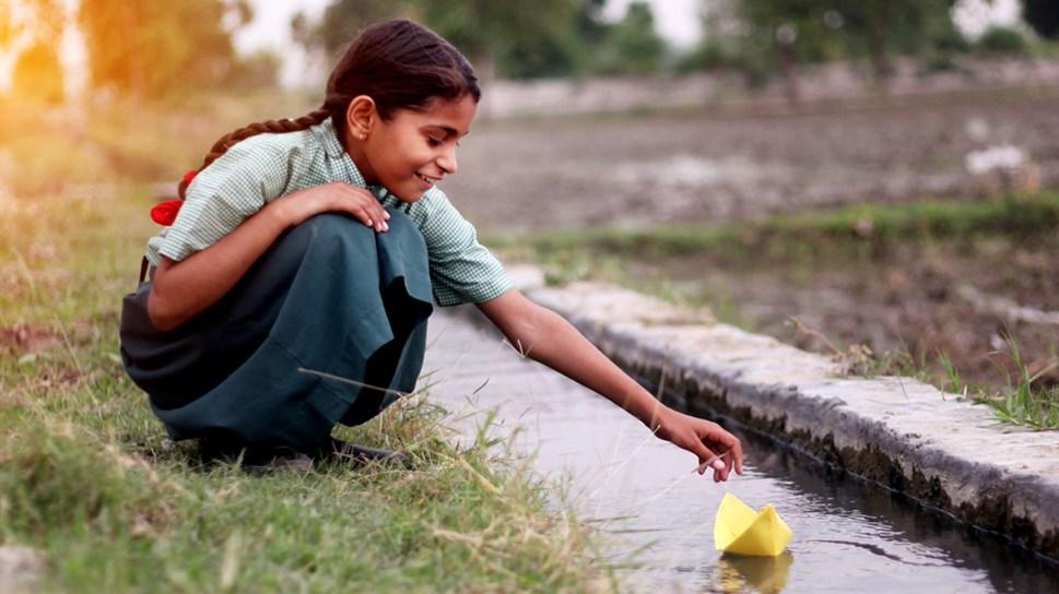 Little girl of elementary age wearing school uniform sitting near water canal outdoor in the field & flowing paper boat in to the water.