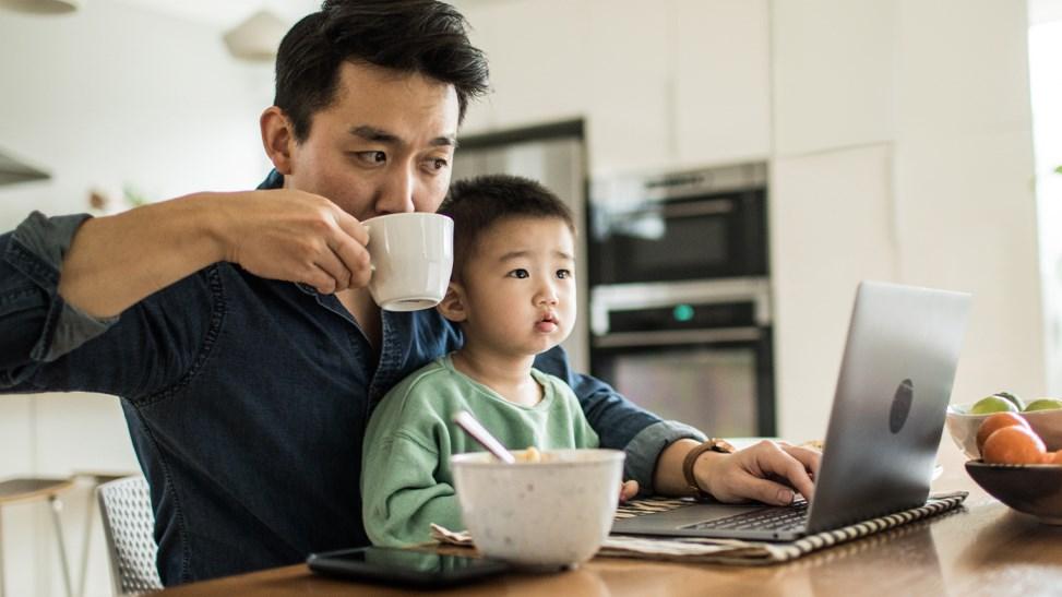 Father multi-tasking with work, with his young son at the kitchen table.