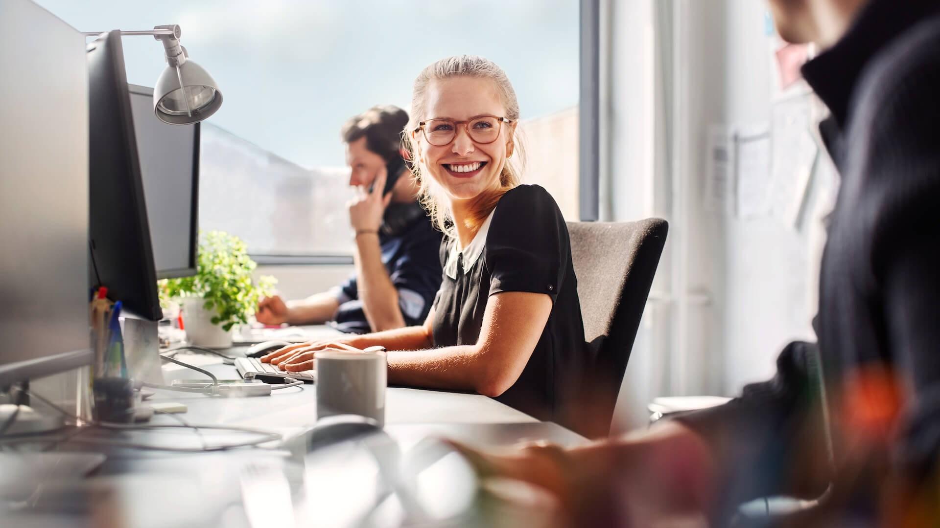 A woman sits in front of a computer in an office environment. She smiles in conversation with her colleague. The man next to her is on the phone.