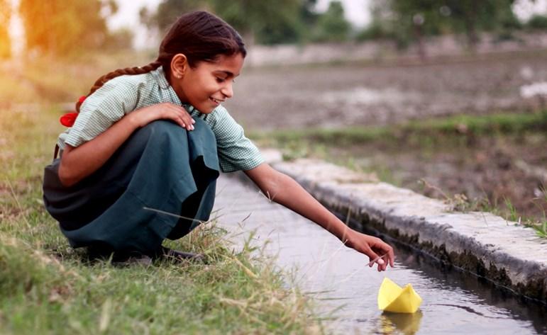 Little girl of elementary age wearing school uniform sitting near water canal outdoor in the field & flowing paper boat in to the water.