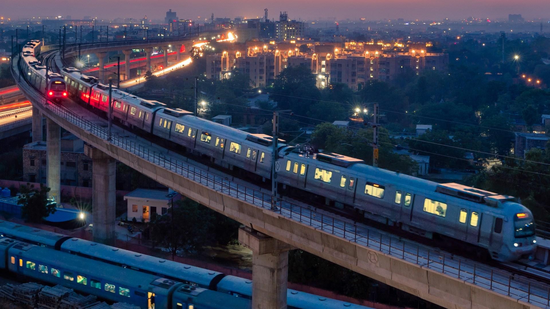 Two trains from the Jaipur Metro leading into a lit up city.